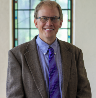 A man wearing glasses, a brown blazer, and a purple tie with a patterned design smiles at the camera. He is standing in front of a window with stained glass details.